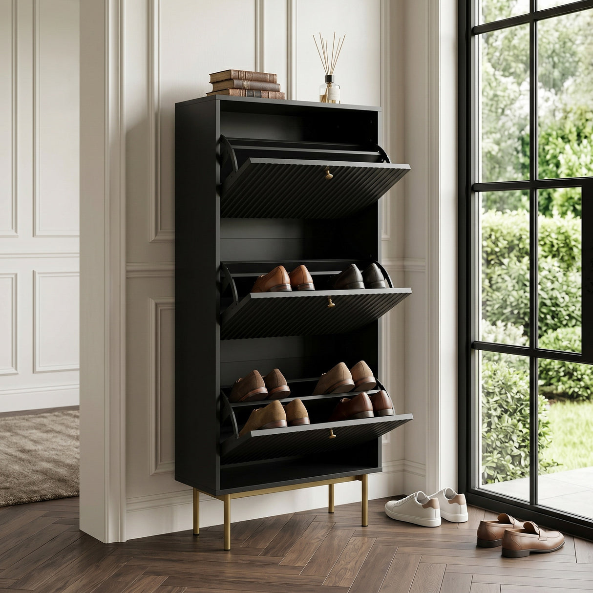 Devon Shoe Cabinet in black with three open tilt out compartments, gold legs and handles, styled with shoes beside a large window.