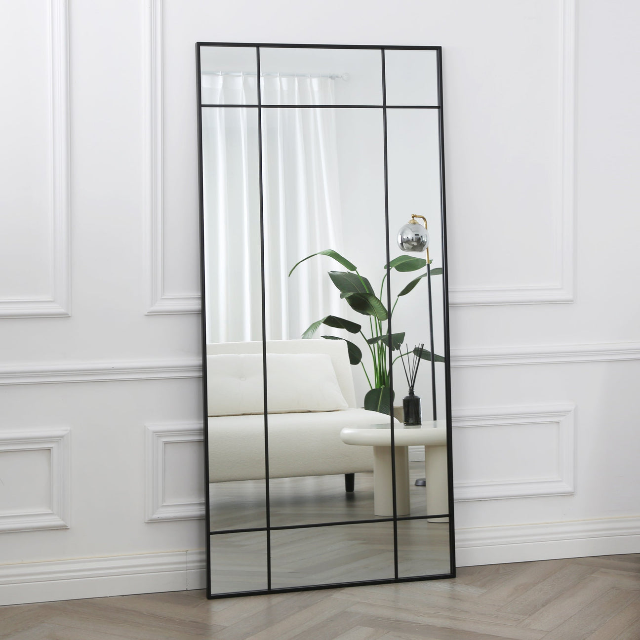 A large industrial style mirror with a black frame, standing in a room with light decor, providing a full-length reflection.