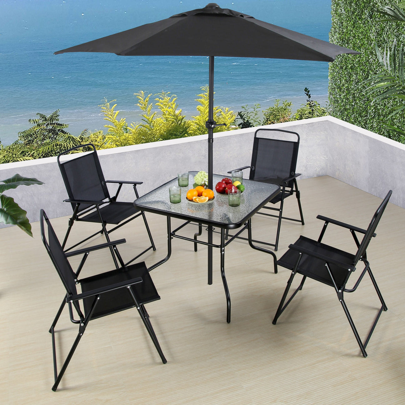 A patio dining set comprising a glass topped table, four foldable chairs, and a black umbrella, placed on a tiled patio with a scenic background.