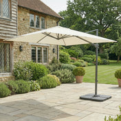 Beige patio umbrella on a stone patio in front of a stone building with greenery.