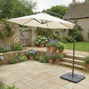 Beige patio umbrella on a stand in a garden setting with flowers and a stone building.